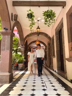 a man and woman are walking down an archway way with potted plants hanging from the ceiling