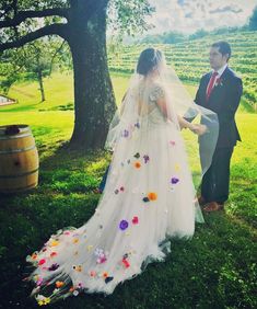 a bride and groom standing in the grass with flowers on their wedding dress, looking at each other