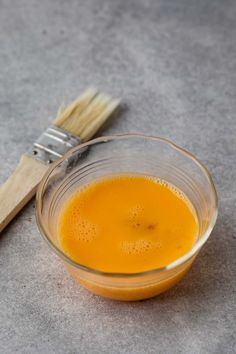 a glass bowl filled with orange liquid next to a wooden toothbrush on a gray surface