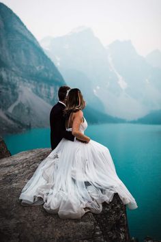 a bride and groom sitting on the edge of a cliff looking out over a lake