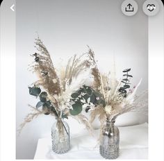 two vases filled with dried flowers on top of a white tablecloth covered table