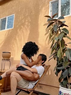 two people sitting on a wooden chair in front of a house with potted plants