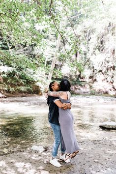 two women hugging each other in front of a river with trees and rocks behind them