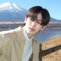 a young man wearing a tan jacket and tie standing in front of a lake with a mountain in the background