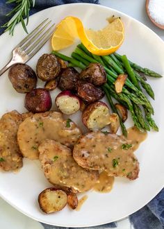 a white plate topped with meat covered in gravy next to asparagus and potatoes