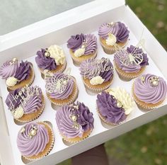 a box filled with purple cupcakes covered in frosted icing on top of a table