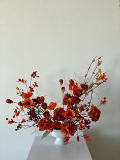 a white vase filled with lots of red flowers on top of a wooden table next to a wall