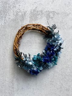 a wicker wreath with blue flowers and greenery on the outside, against a concrete background