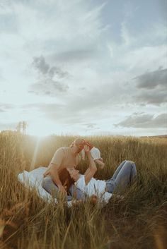 a man and woman laying on the ground in tall grass with sun shining through clouds