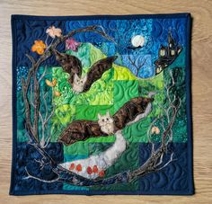 a quilted wall hanging with cats and bats in the night sky, on top of a wooden table