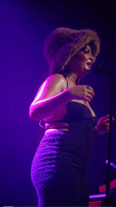 a woman standing in front of a microphone with her hands on her hips and wearing a purple dress