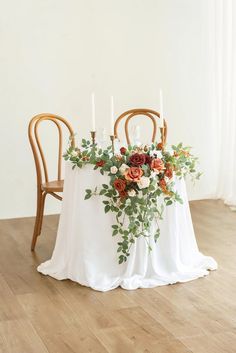 an arrangement of flowers and candles on a white table cloth in front of two wooden chairs