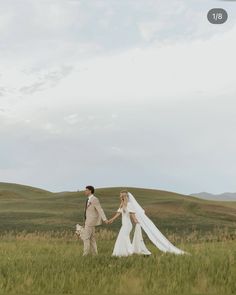 a bride and groom holding hands walking through the grass with mountains in the background on their wedding day