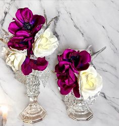 two silver vases with purple and white flowers on a marble counter top next to candles