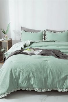 a bed with green sheets and pillows in a white room next to a plant on the floor