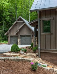 two garages in front of a house surrounded by trees and rocks with flowers on the ground