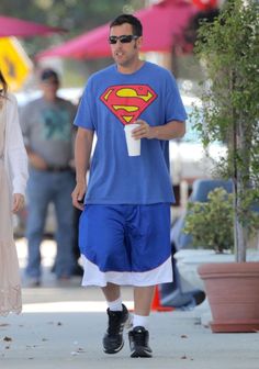 a man in a superman t - shirt and shorts walks down the street with a woman