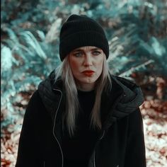 a woman with long blonde hair wearing a black hat and jacket in the woods looking at the camera