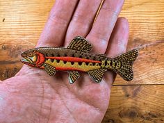 a hand holding a small brown and orange fish on it's left side, next to a wooden table