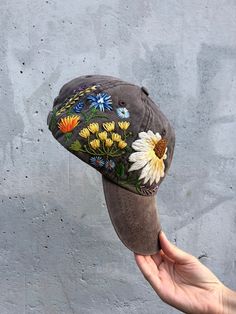 a person holding up a hat with flowers on the front and side, against a concrete wall