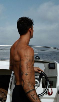 a man with tattoos on his back standing in front of a white boat and holding a steering wheel