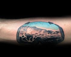 a man's arm with a snow goggles tattoo on his left forearm and an image of mountains in the background