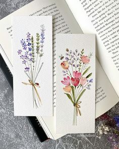 three cards with watercolor flowers on them sitting next to an open book and a pen