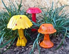 three colorful mushrooms sitting on the ground next to grass and flowers in front of a stone wall