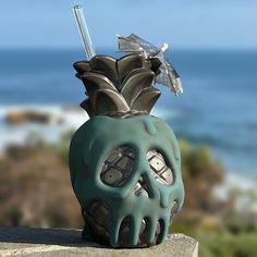 a skull shaped vase with a toothbrush sticking out of it's mouth, sitting on a ledge near the ocean