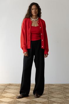 a woman standing in front of a white wall wearing black pants and a red cardigan