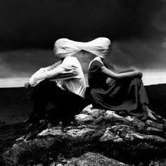 two people sitting on top of a rocky hill under a cloudy sky with their heads wrapped around each other