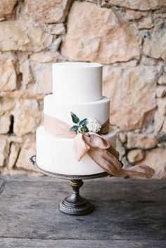 a three tiered white wedding cake with pink ribbon and flowers on top, sitting on a table in front of a stone wall