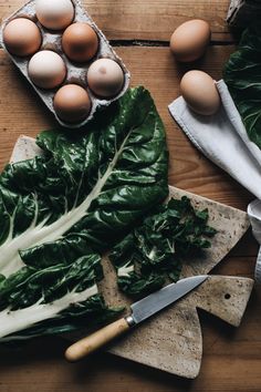 some greens and eggs are on a table next to a cutting board with a knife