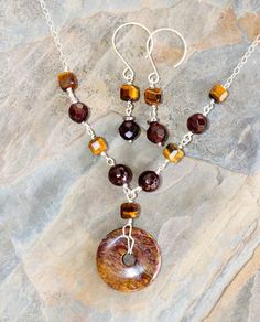 This tiger eye jewelry set is perfect for fall! This natural stone necklace features a natural pietersite donut bead.  It measures 1 inch in diameter.  The stone is various shades of golden brown with natural inclusions.  I wire wrapped the petersite bead with sterling silver wire, which I bent for added design interest.  I accented this pietersite, garnet and tiger eye necklace with natural stone beads:  golden brown, cube shaped tiger eye and burgundy red garnet.  There are a total of five tiger eye beads and four garnet beads.  This pietersite, tiger eye, and garnet necklace is finished with a sterling silver chain. Length:  18 in.; pendant hangs 1.5 in. below See the 7th photo for scale. The matching earrings feature natural stone, tiger eye beads. They are various shades of golden bro Round Amber Gemstone Beads Jewelry, Brown Jewelry With Polished Round Beads, Brown Natural Stones Round Jewelry, Brown Gemstone Round Pendant Jewelry, Brown Round Gemstone Beads Jewelry, Brown Gemstone Jewelry With Round Beads, Garnet Jewelry Set, Tiger Eye Earrings, Autumn Necklace