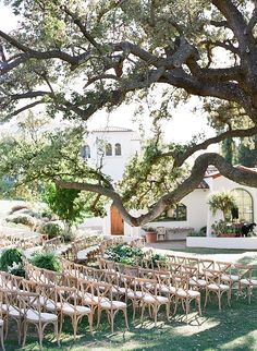 an outdoor ceremony set up under a large tree in front of a white house with lots of chairs