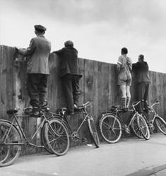 several men standing on bicycles in front of a wooden fence and looking at the sky