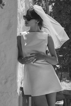 a woman in a short white dress leaning against a wall with her veil over her head