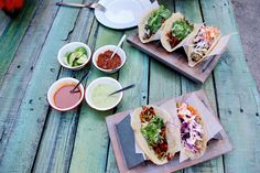 three tacos are sitting on wooden trays with dipping sauces and condiments