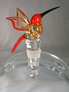 a glass humming bird sitting on top of a clear stand with a red and yellow flower