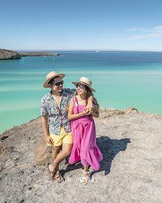 a man and woman posing for a photo on the rocks by the water with their arms around each other