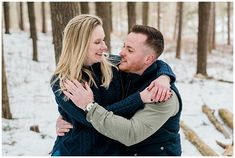 a man and woman hugging each other in the woods with snow on the ground behind them