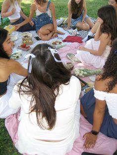 a group of women sitting on top of a pink blanket eating food at a park