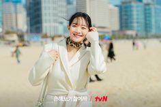 a woman in white coat standing on beach next to tall buildings and smiling at the camera