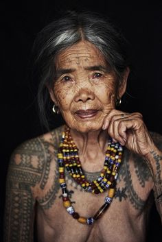 an old woman with tattoos and beads on her chest is posing for the camera in front of a black background