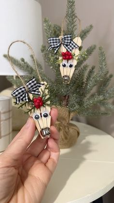 a hand holding two wooden reindeer ornaments in front of a christmas tree with red berries on it