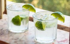 two glasses filled with ice and limes sitting on a marble table next to each other