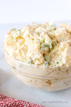 a glass bowl filled with potato salad on top of a white tablecloth next to a red and white napkin