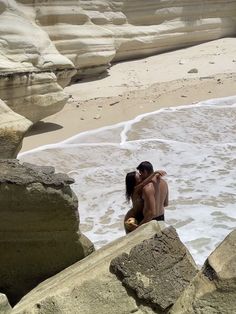 two people sitting on rocks in the water at the beach, one holding his arm around the other's neck