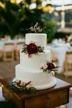 a white wedding cake with red flowers and greenery sits on a wooden table in front of candles