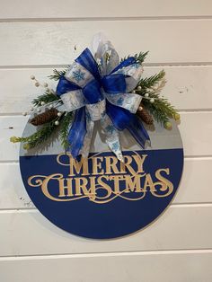 a merry christmas sign hanging on the side of a white wall with blue ribbon and pine cones